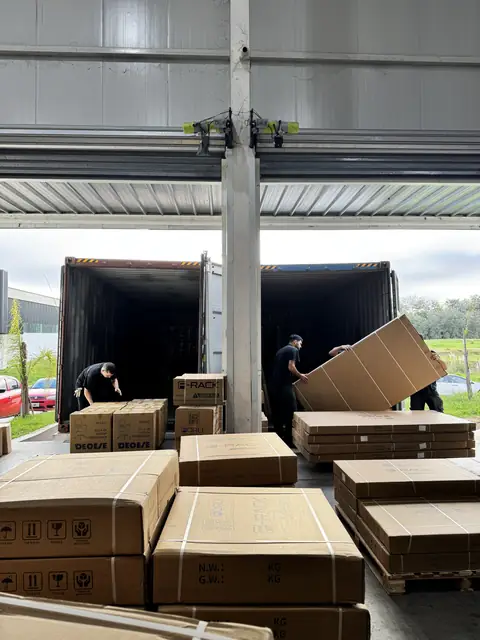Dos operarios de LMC bajan del contenedor una caja de escritorio más grande que ellos, junto a pallets de cajas largas